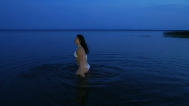 Молодая женщина в белом купальнике в озере делает всплеск на воде в сумерках . — стоковое видео