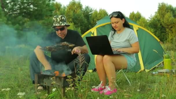 Mężczyzna i kobieta są w pobliżu namiotu. Kobieta pracuje na laptopie, człowiek jest mieszając węgle w grill i trzyma w ręku mięso z grilla. — Wideo stockowe