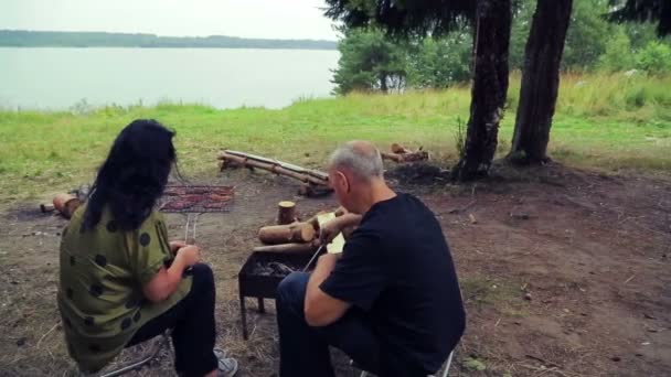 Чоловік і жінка на березі річки сидять біля барбекю і готують на грилі рибу . — стокове відео
