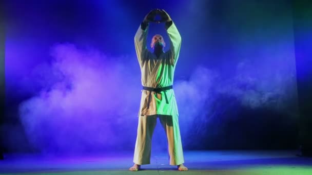 Человек в белом кимоно занимается карате на фоне цветного дыма — стоковое видео