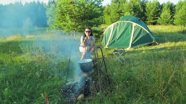 A young woman sits by the fire near the tent, watches the fire, over which the bowler hanging and drinking tea. — Stock Video