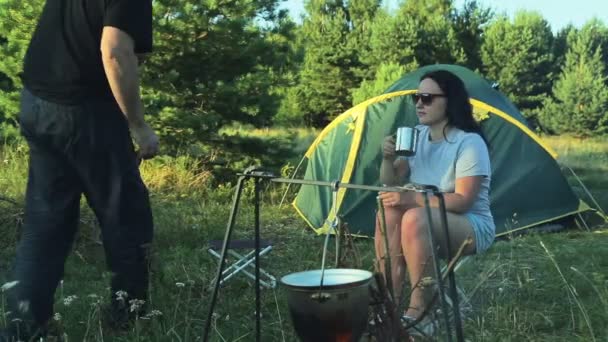 A man and a woman in sunglasses are sitting by the tent, drinking tea and watching the pot hanging over the fire. — Stock Video