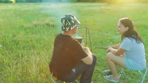 Mężczyzna i kobieta siedzą przy ognisku, nad którym wisi melonik. Strzelanie z tyłu. — Wideo stockowe