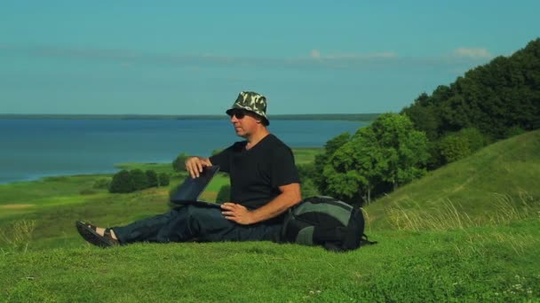 Un hombre con una mochila sentado en el borde de la montaña con vistas al lago abre una computadora portátil . — Vídeos de Stock