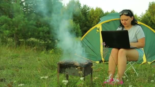 Een man en een vrouw zijn zit in de buurt van een tent en grillen met het grillen van vlees. Vrouw die op laptop werkt. — Stockvideo