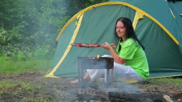 Žena v zeleném tričku sedí v lese na pozadí stanu, příprava večeře na její pánev. Celkový plán
