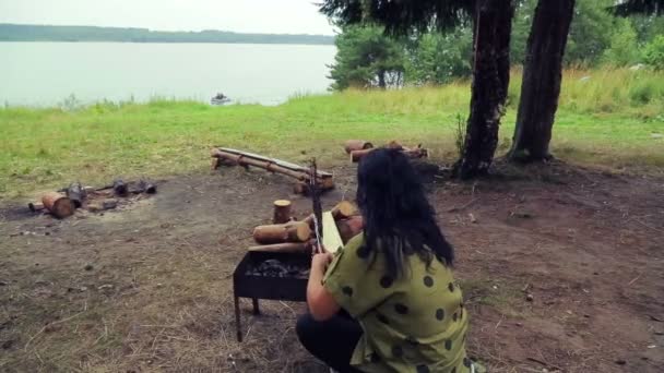 Una mujer en la orilla del río se sienta en la barbacoa y gira la parrilla con un pescado . — Vídeos de Stock