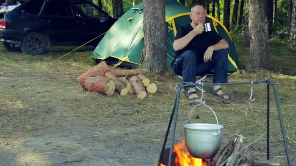 Мужчина Пьет Чай Возле Палатки Смотрит Горшок Висящий Над Огнем — стоковое видео