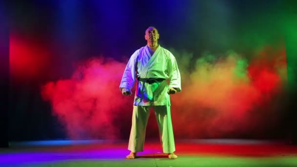 Un hombre muestra golpes de karate sobre un fondo negro con humo de color — Vídeos de Stock