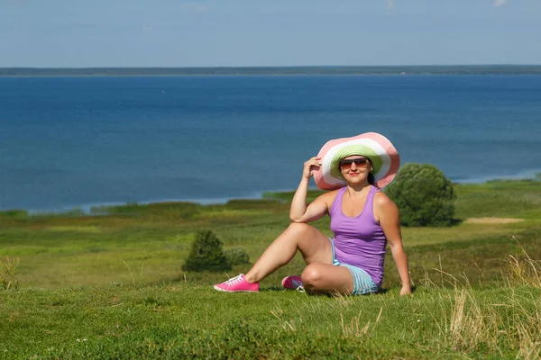 Joven mujer feliz en sombrero y gafas de sol está sentado en una colina frente a un lago . —  Fotos de Stock