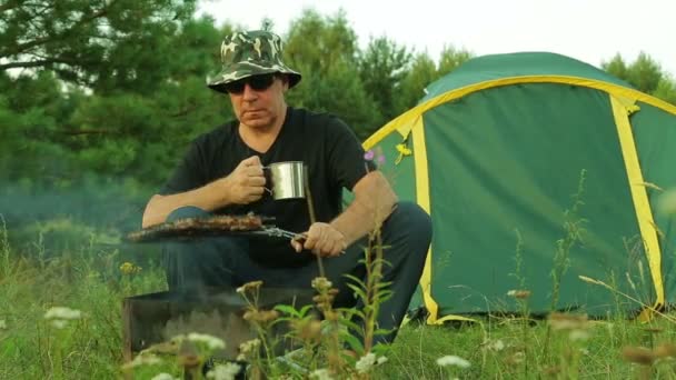 Um homem senta-se junto à tenda, bebe chá e transforma a carne na grelha . — Vídeo de Stock