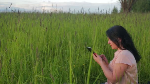 Žena sedí v zelené trávě a pracuje na tabletu v parku mezi plán. Kamera se pohybuje na dolly — Stock video