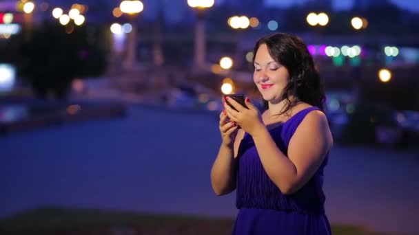 Hora de la noche una mujer joven en un vestido azul en la calle leyendo un mensaje en un teléfono inteligente está sonriendo y soñando mirando hacia arriba — Vídeos de Stock