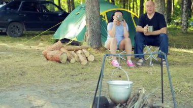 Turistler, bir erkek ve bir kadın çadırda oturan, çay içme ve ateşin üzerinde asılı Melon izlerken.
