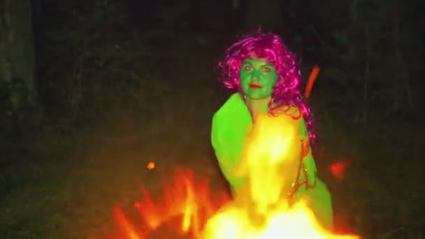 The green witch is warmed by the fire at night in the forest. — Stock Video