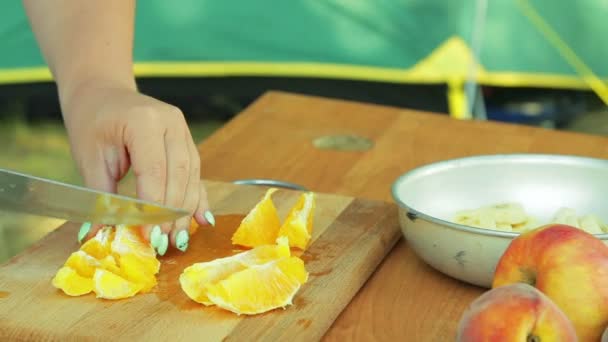 A young woman is knifing an orange on a wooden board for fruit salad. — Stock Video