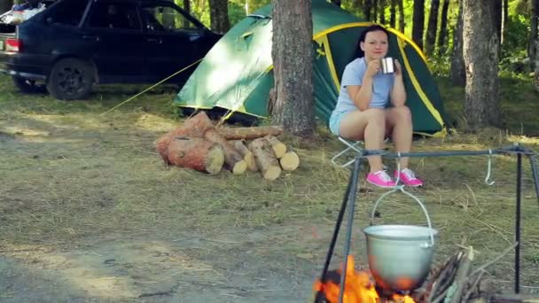 Молодая женщина пьет чай возле палатки и смотрит на боулер, висящий над огнем . — стоковое видео