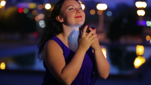 Вечірній час молода жінка в блакитній сукні спілкується в соціальних мережах на смартфоні на тлі вуличних ліхтарів, розмитих в стороні. Панорама зліва направо . — стокове відео