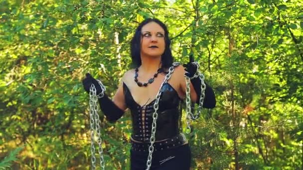 A woman is a sorceress in black clothes with a chain in her hands in the forest. Halloween. — Stock Video