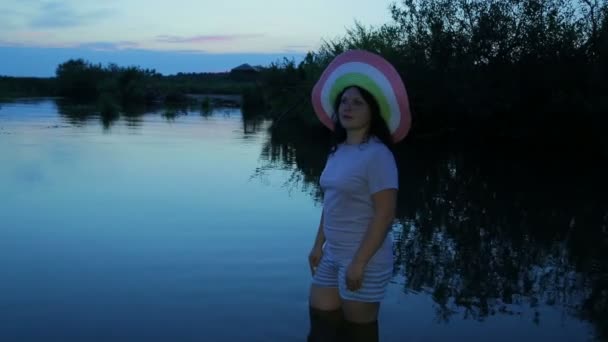 Chica joven con sombrero en el lago en la rodilla. Crepúsculo — Vídeos de Stock
