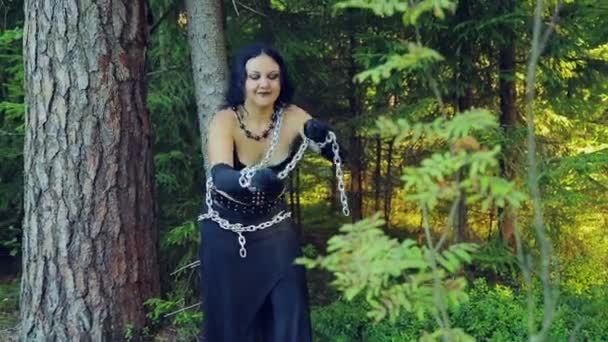 Une femme en colère, une sorcière vêtue de noir dans une forêt attachée avec des chaînes à un arbre, tente de se libérer. Halloween. Style gothick . — Video