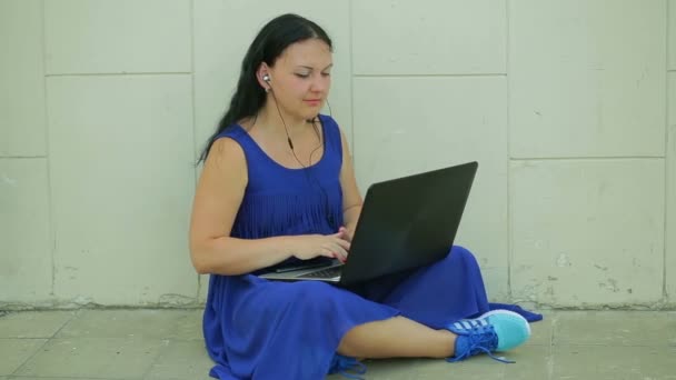 A female student is working on a laptop in the street next to a white wall. Panorama from left to right — Stock Video