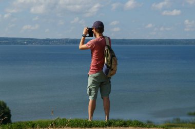 Onun arkasında bir sırt çantası ile bir gezgin bir tepe üzerinde duran ve göl bir resim çekmek.