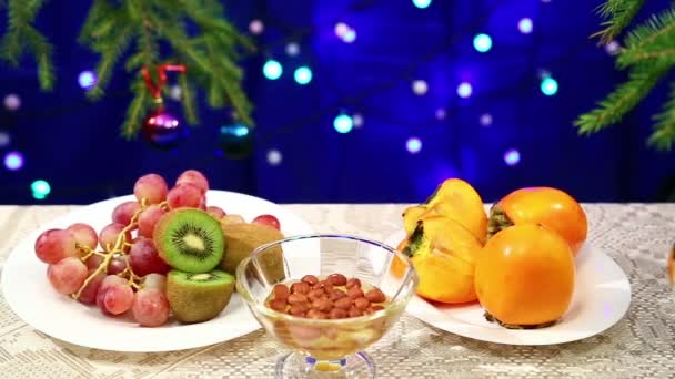 Pratos com kiwi, uvas e nozes com mel e estão sobre a mesa com bolas decoradas com bolas de Natal e luzes bokeh borradas — Vídeo de Stock