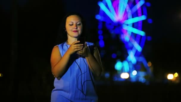 A young woman on the quay in headphones reads news on the Internet on the phone against the backdrop of a glowing ferris wheel. — Stock Video