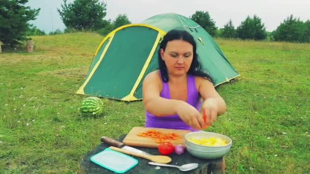 Une jeune femme s'assoit près d'une tente et empile des tranches de carotte hachée dans une assiette avec des légumes . — Video