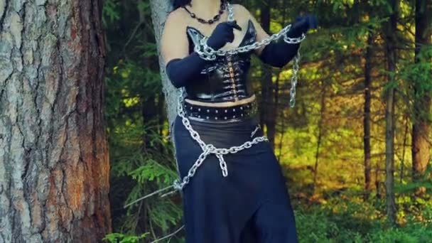 Een vrouw van een heks in zwarte kleding in een forest is verbonden met kettingen aan een boom en probeert om zichzelf vrij. Halloween. Gothick stijl. — Stockvideo