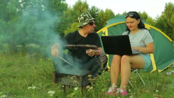 Ein Mann und eine Frau sind draußen und grillen Fleisch auf dem Grill. Frau arbeitet am Laptop. — Stockvideo