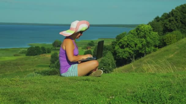 Een vrouw in een stro hoed zit op een berg met uitzicht op het meer en de laptop deksel sluit. — Stockvideo