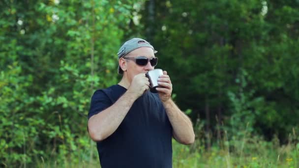 En man står på kanten av skogen dricka te från en mugg och beundra naturen. — Stockvideo