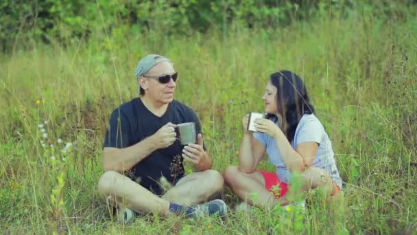 男と女の観光客が草の上に座って、マグカップからお茶を飲むと話して. — ストック動画
