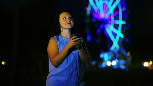 Veselá mladá žena na nábřeží ve sluchátkách poslouchat hudbu a tanec na pozadí zářící ruské kolo. — Stock video