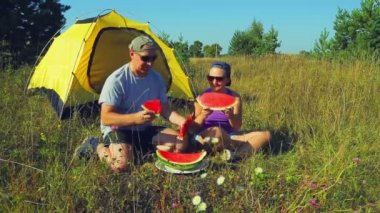 Çadırın yakınında bir açıklıkta genç bir kadın elinde bir bıçakla bir karpuz yanında oturur.