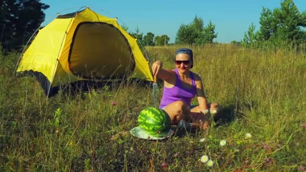 A young woman in a clearing near the tent sits next to a watermelon holding a knife. — Stock Video