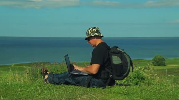 A man with a backpack behind his back sits on the edge of the mountain and prints on a laptop. Shot from the side. — Stock Video