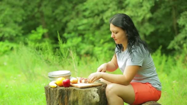 Jonge vrouw slicks stukjes perzik voor salade op een houten plank op de rand van een bos. — Stockvideo