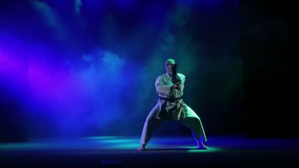 Человек в кимоно занимается карате - выполняет упражнения на фоне цветного дыма — стоковое видео