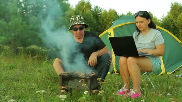 一个男人和一个女人坐在帐篷附近。一个女人正在用笔记本电脑, 一个男人在烤烧烤上看肉烧烤。. — 图库视频影像