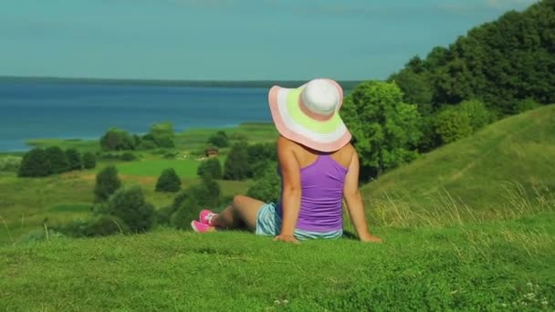 戴着帽子的女人坐在山上欣赏湖景。从后面射击. — 图库视频影像