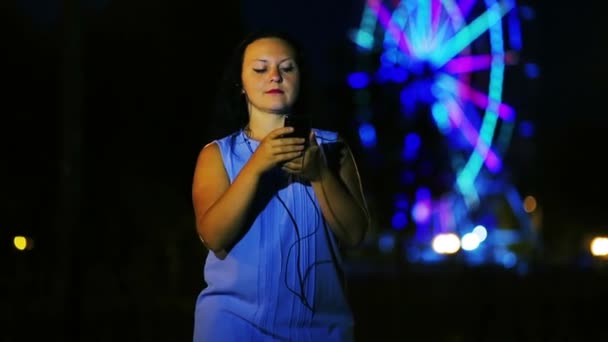 Una mujer joven en el muelle en los auriculares escribe un mensaje en el teléfono en el telón de fondo de una rueda de la fortuna brillante . — Vídeos de Stock