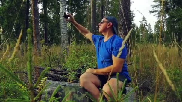 Güneş gözlüklü erkek bir turistik ve beyzbol şapkası ormandaki düşmüş bir ağaç üzerinde oturur ve telefonda fotoğraf çeker. — Stok video
