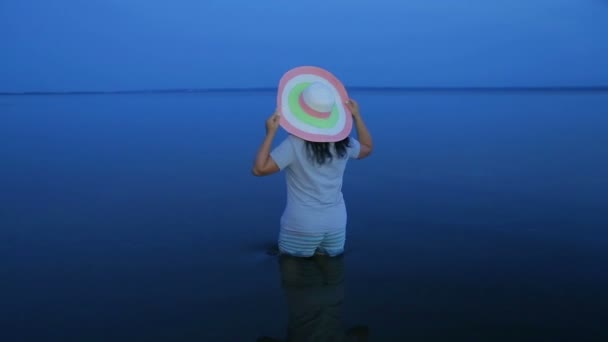 Uma jovem mulher sorridente em shorts e um chapéu em pé junto ao joelho em um lago e admirando o pôr do sol . — Vídeo de Stock