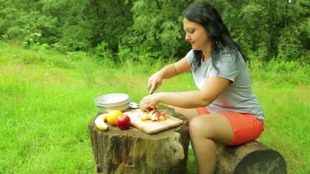 Jonge vrouw toeristische is zittend op een boomstronk aan de rand van een bos en snijden stukjes perzik voor salade op een houten bord. — Stockvideo