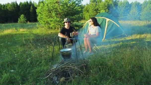Чоловік і жінка туристи сидять біля багаття і п'ють гарячий чай . — стокове відео