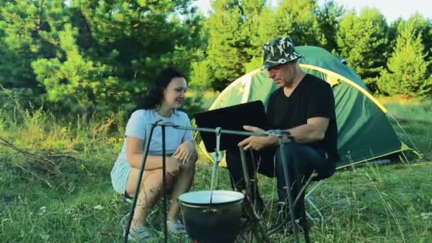 Um homem e uma mulher estão sentados em uma tenda, observando um jogador pendurado sobre um fogo e trabalhando com um laptop . — Vídeo de Stock