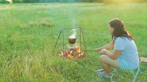 La donna siede vicino al fuoco, sopra il quale la bombetta appesa e mescolando carboni. Girato da un lato . — Video Stock